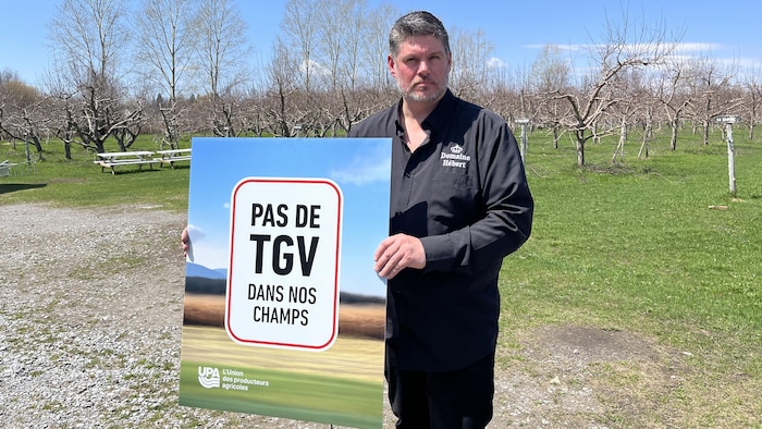 Près de ses pommiers, Christian Hébert tient une affiche montrant un champ et un panneau "pas de TGV dans nos champs". 