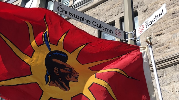 Un drapeau warrior flotte au vent sous un panneau de rue au nom de Christophe Colomb à Montréal.