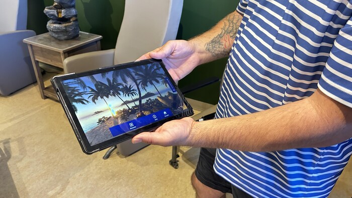 Un homme tient une tablette sur laquelle on observe une plage et des palmiers.