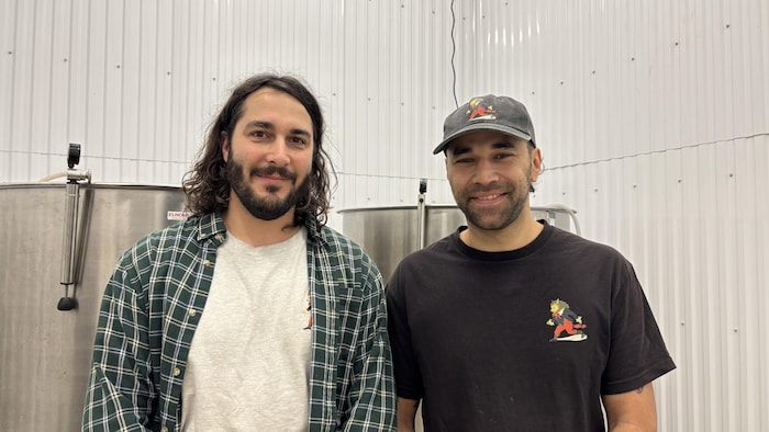 Un portrait de deux hommes souriants, se tenant côte à côte devant deux cuves de fermentation industrielles en acier inoxydable. L'homme de gauche a les cheveux longs et porte une chemise à carreaux. L'homme de droite porte une casquette et un t-shirt noir.