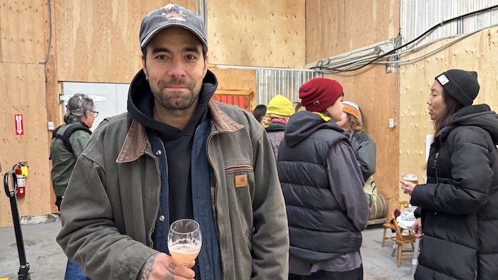 Un homme à la barbe courte, portant une casquette, une veste de travail kaki et des tatouages à la main, tient un verre de cidre rosé. Il se tient au premier plan d'un intérieur aux murs de bois et de tôle, avec d'autres personnes en vêtements d'hiver derrière lui.
