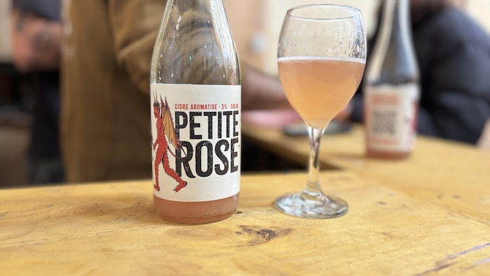 Un gros plan sur une bouteille de cidre rosé nommée "PETITE ROSE" et un verre à pied rempli du même cidre, posés sur une table en bois. L'arrière-plan flou montre des personnes.