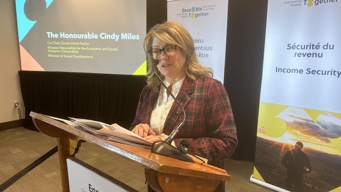 Une femme parle dans un micro, un écran et des bannières derrière elle.