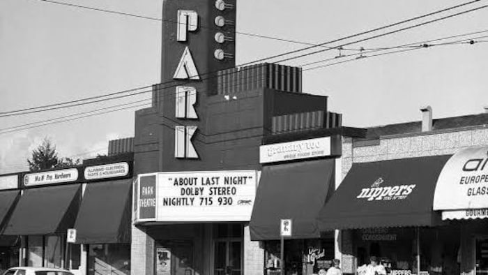 Des commerces et un cinéma dans une rue où passe une voiture. 