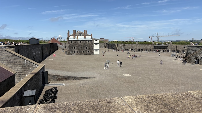 Sous un ciel bleu et clair, des touristes déambulent sur un grand espace entouré de hautes fortifications.