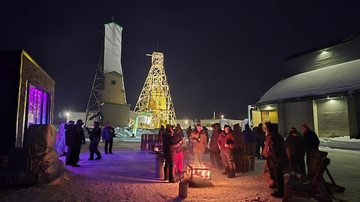 Des gens sont réunis autour d'un feu en plein coeur de la Cité de l'Or, avec des chevalements de mine éclairés.