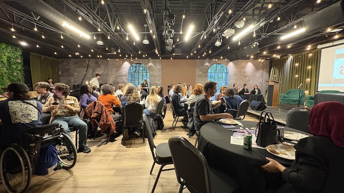 La salle est remplie de personnes assises autour de tables rondes.