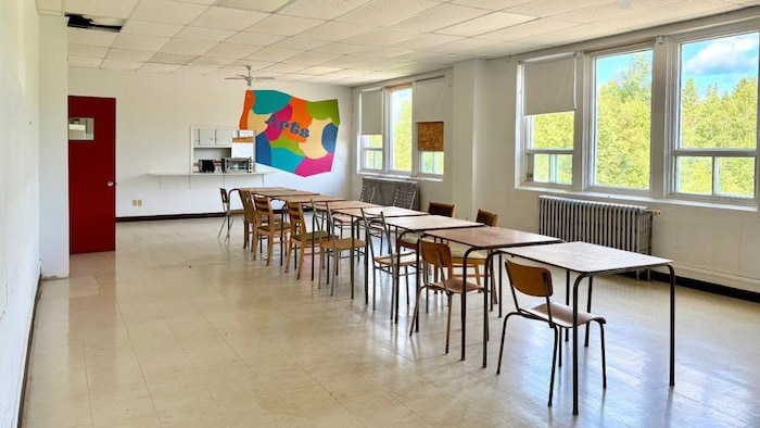 Des bureaux sont installés de bout en bout dans une classe autrement vide. Un peinture sur le mur indique que c'était une classe d'arts.