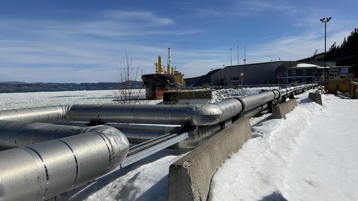 Un gros tuyau gris en avant-plan avec un bateau en arrière-plan au Port de Saguenay.