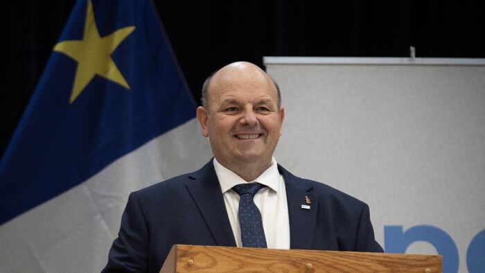 Claude Bourgeois debout, derrière un pupitre de conférence. Un drapeau acadien est derrière lui.