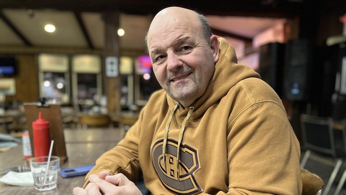 Un homme avec un chandail à capuche jaune du Canadien de Montréal est assis à une table en bois dans un bar, les deux mains croisées.