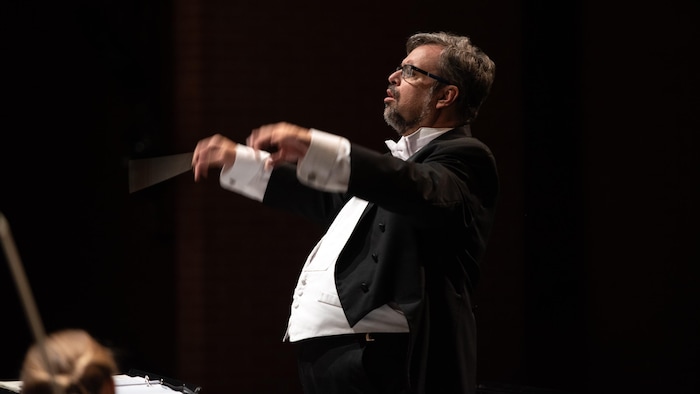 Baguette de chef d'orchestre à la main et les bras levés, Claude Lapalme dirige des musiciens sur scène.