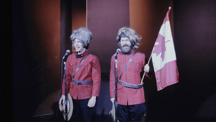 Claude Meunier et Jacques Grisé sur scène habillés en policier de la gendarmerie royale du Canada. Jacques Grisé tient un drapeau du Canada. 