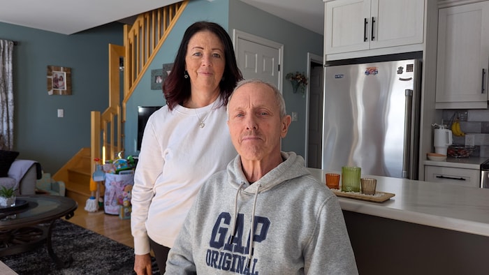 Un homme assis, une femme debout derrière lui, dans une maison, avec un frigidaire chromé et luisant derrière eux.