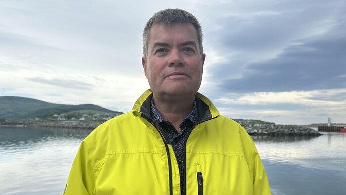 Claudio Bernatchez est vêtu d'un imperméable et se tient debout devant le port de Gaspé.