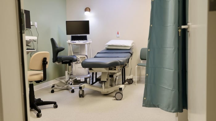 Une table d'examen médical, deux chaises de bureau à roulettes et un écran d'ordinateur dans une petite pièce qui se ferme avec un rideau.