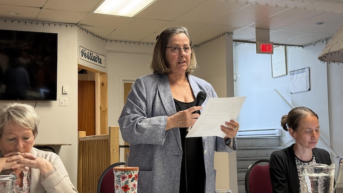 Josseline Lepage parle aux citoyens, un micro et une feuille de papier dans les mains. Lyne Dupras et Sarah Charbonneau sont assises à ses côtés.