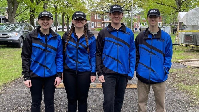 Juliette Gagné, Raphaëlle Jourdain, Charles-Antoine Massé et Steve Jean posent pour la caméra, en rang sur un terrain de pétanque. Ils portent des casquettes et des chandails à l'effigie du Club Bouliste de Baie-Comeau.