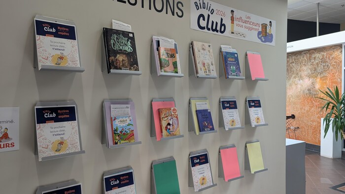 Un mur beige accueille plusieurs présentoirs métalliques alignés proposant des livres jeunesse et des brochures promotionnelles.