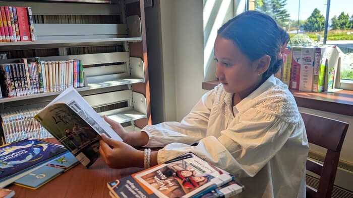 Une jeune fille portant un haut blanc brodé est assise à une table et lit attentivement un livre ouvert.