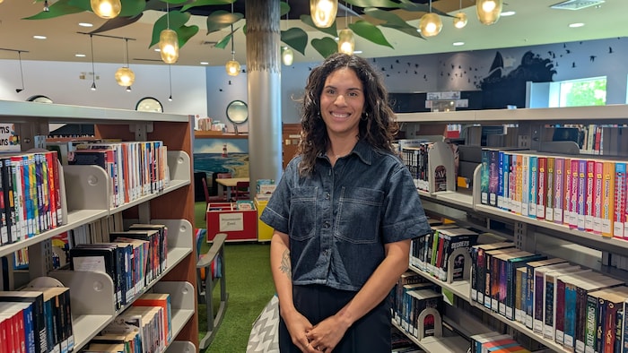 Une femme aux cheveux ondulés et vêtue d'une chemise en jean pose en souriant dans une bibliothèque.