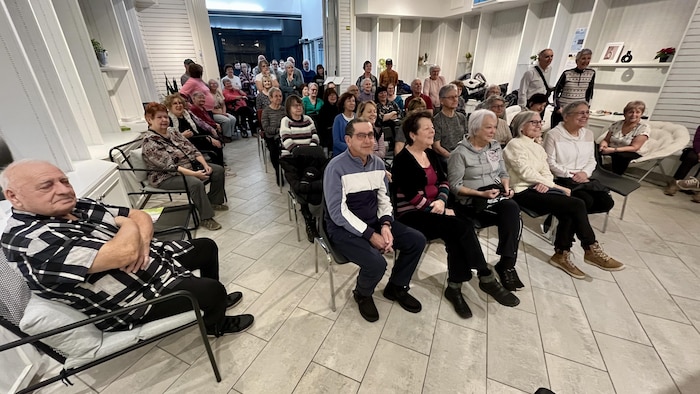Une salle d'un centre commercial est remplie de personnes assises sur des chaises, et d'autres debout à l'arrière.
