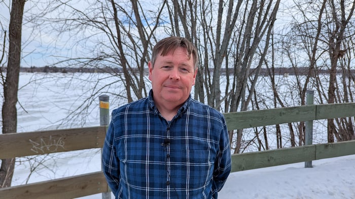 Un homme en chemise à carreaux bleue, sans manteau d'hiver, est debout les bras derrière le dos, tourné à un fleuve gelé.