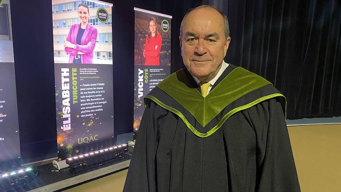 Un homme d'un certain âge pose en toge noire ornée d'un collet vert mousse. Derrière lui, des bannières promotionnelles de l'UQAC présentent des diplômées sur une scène éclairée par des projecteurs au sol.