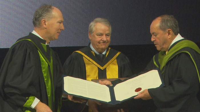 Jean-François Lépine reçoit officiellement son diplôme des mains de deux dignitaires universitaires lors d'une cérémonie de l'UQAC. Les trois hommes portent des toges académiques et le document, orné d'un sceau rouge, est présenté ouvert devant eux.