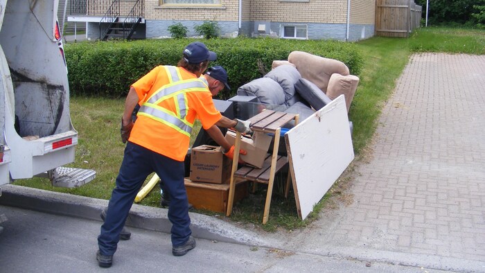 Deux employés ramassent de gros déchets sur le bord de la rue.