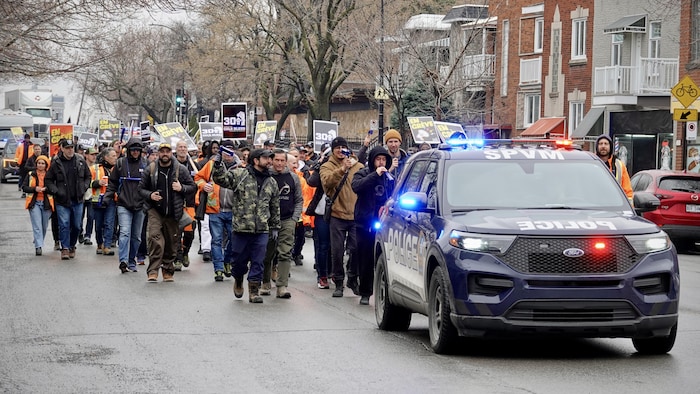 Des grévistes des cols bleus manifestent, accompagnés d'une voiture de police sur une rue de Montréal.