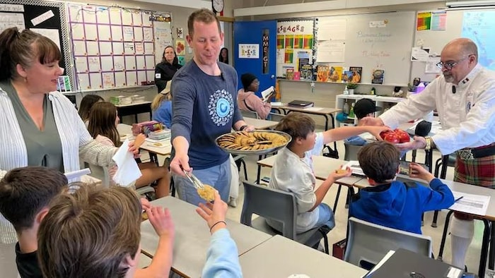 Deux hommes qui distribuent des biscuits et des pommes à des élèves dans une salle de classe.