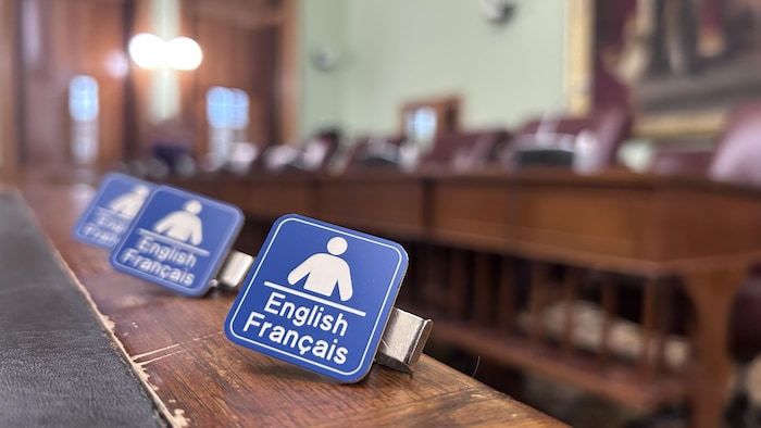 Une photo de la salle des comités parlementaires de l'Assemblée législative. En premier plan, il y a trois broches signalant que des services sont disponibles en français et en anglais.