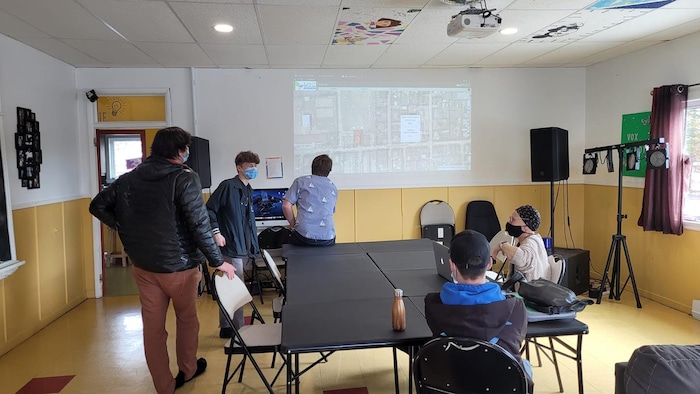 Un plan est projeté sur un mur, et un groupe de personnes sont autour d'une table de réunion, ils portent des masque chirurgicaux.