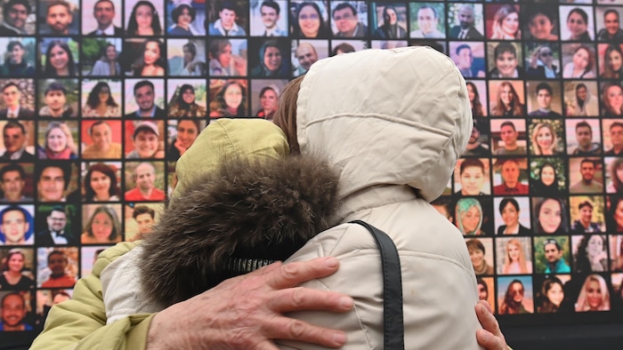 Two women console each other with portraits of victims of the crash of flight PS752 in the background.