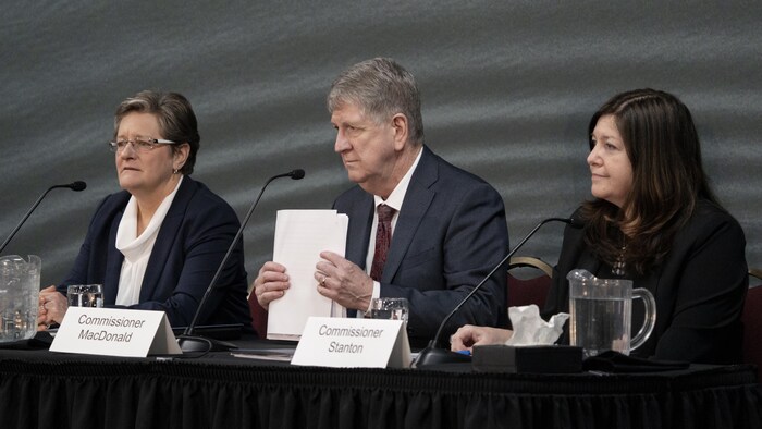 Leanne Fitch, Michael MacDonald et Kim Stanton assis à une longue table, des micros devant eux. Michael MacDonald tient une pile de documents.