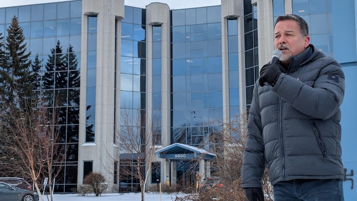 Larry Imbeault parle dans un micro devant un bâtiment.