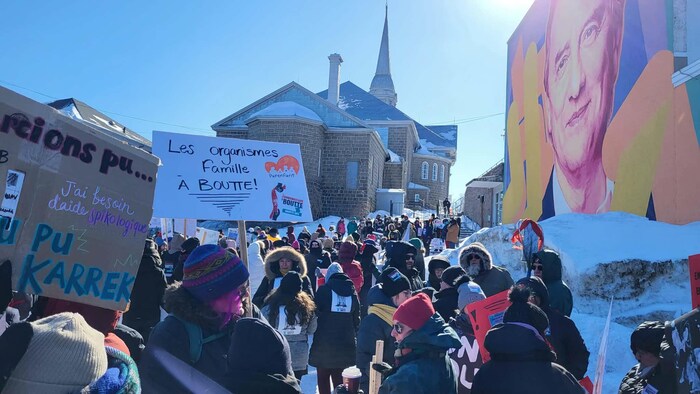 Une large foule de manifestants, vue de dos, se dirige vers une église en pierre sous un soleil éclatant. Des pancartes artisanales expriment l'épuisement des organismes famille, à côté d'un mur orné d'une grande murale colorée.