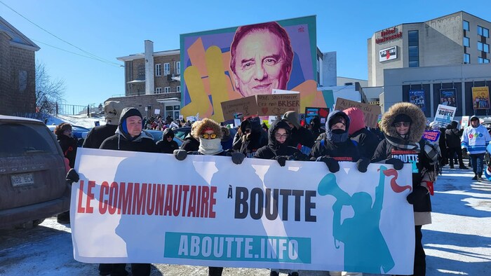 Un groupe de manifestants en tenue d'hiver tient une large bannière indiquant « Le communautaire à boutte » devant une murale colorée représentant un visage. L'action se déroule à l'extérieur, sous un ciel bleu, près de l'Hôtel Universel, à Alma.