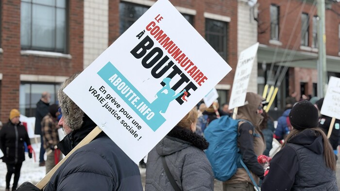 Des gens sont dehors et l’un d’eux tient une pancarte sur laquelle est écrit Le communautaire à boutte, en grève pour une vraie justice sociale.