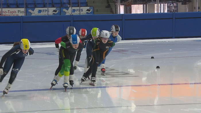 Un groupe de jeunes athlètes patineurs de vitesse en pleine action sur une patinoire intérieure, portant des combinaisons et des casques de différentes couleurs. Ils sont en position de course, penchés vers l'avant sur la glace.
