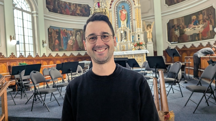 Un homme souriant portant des lunettes et un chandail noir pose au premier plan d'une église.