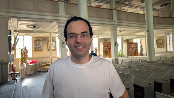 Un homme souriant à lunettes et t-shirt blanc se tient au premier plan à l'intérieur d'une église.