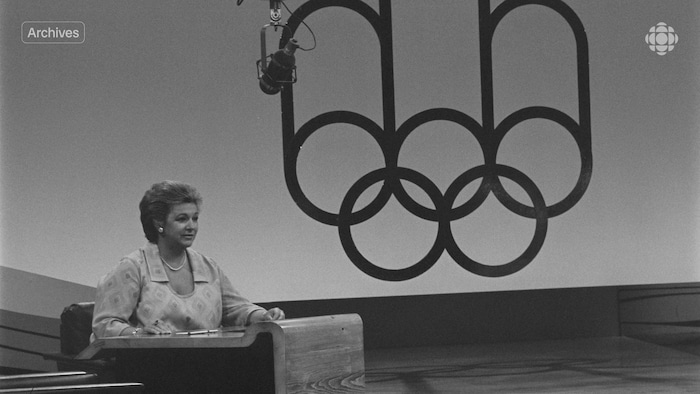 L'animatrice Lise Payette assise à un bureau sur le plateau de son émission « Appelez-moi Lise » durant la période olympique de 1976.