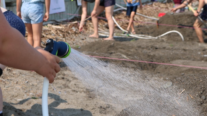 Une personne arrose du sable. Des gens préparent le terrain en arrière-plan. 
