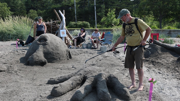 Un homme asperge sa sculpture avec un peu d'eau. Il a construit un cyclope dont la tête et les mains immenses sortent du sable.