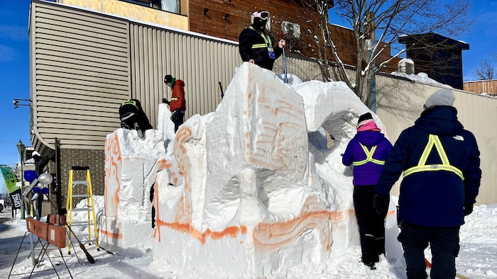 Un bloc de neige couvert de peinture. Des gens se trouvent autour et sur le dessus du bloc pour le sculpter. 
