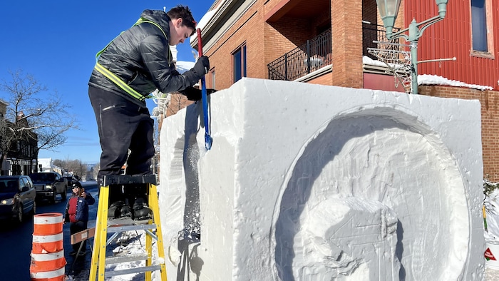 Un étudiant du Cégep de Rivière-du-Loup gratte de la neige pour réduire un des côtés de son bloc de neige. Sur une autre face, un cercle et une flèche dans son centre y sont déjà sculptés.
