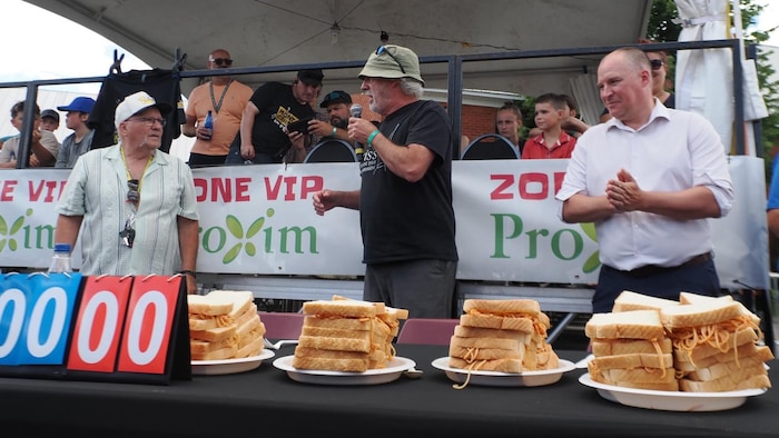 Deux compétiteurs et un animateur devant des sandwichs remplis de spaghetti.