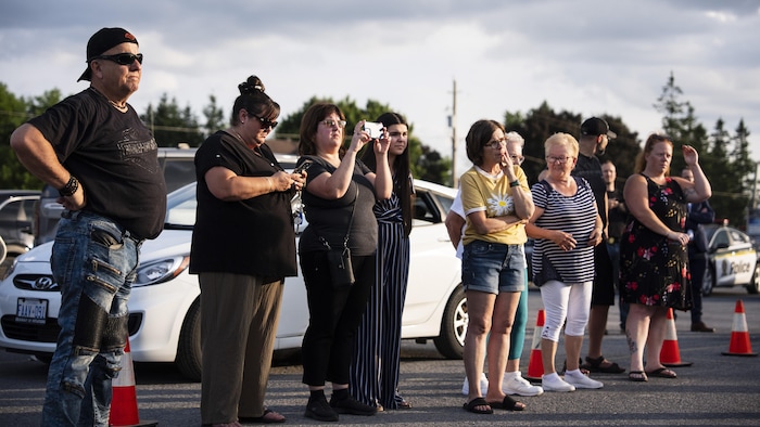 Une dizaines de badauds debout dans un stationnement avec une voiture de police non loin.